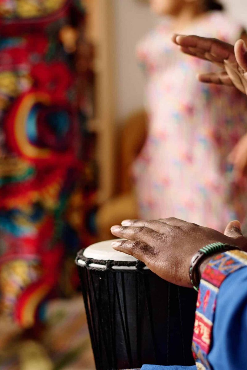 Focus on hands of young African American man in blue national costume beating drum while sitting in front of his wife and daughters dancing